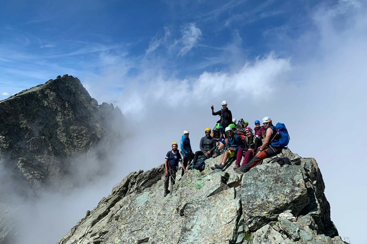 Un gruppo di escursionisti sulla cima di un monte, dietro le nuvole si alzano verso il cielo azzurro, nascondendo in parte un'altra cima