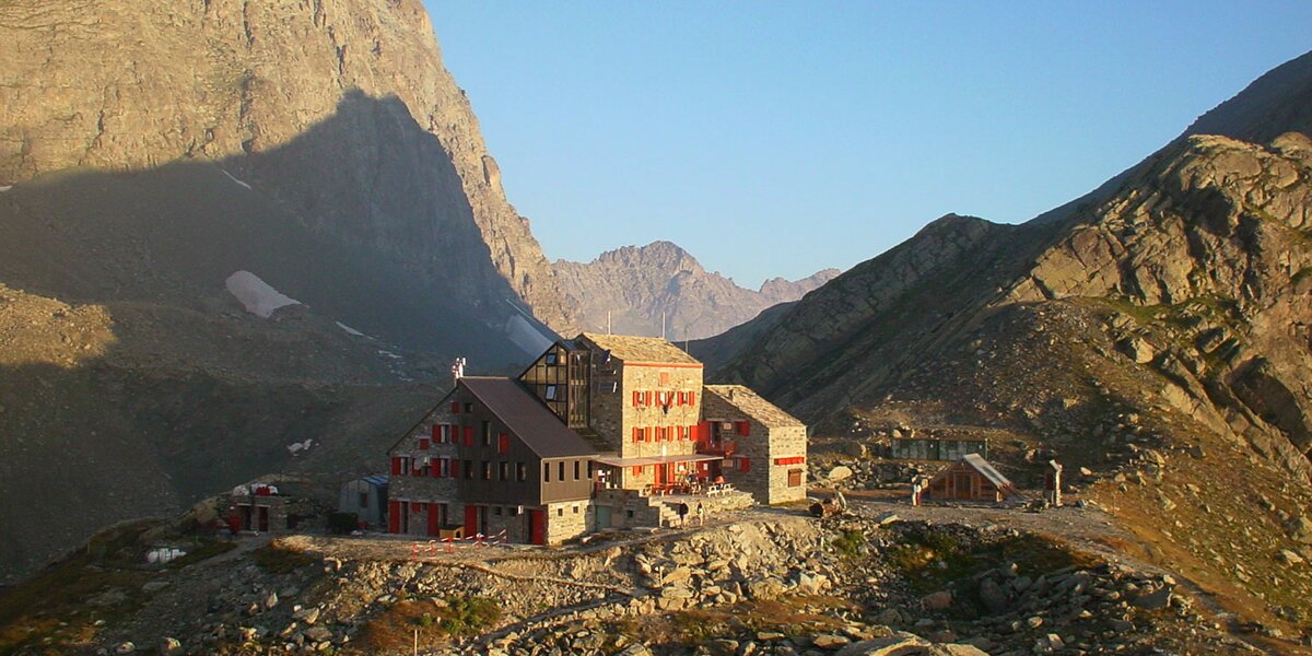 Foto del Rifugio Quintino Sella al Monviso: un edificio in tre corpi con imposte rosse, alle cui spalle si trova la mole del Monviso.