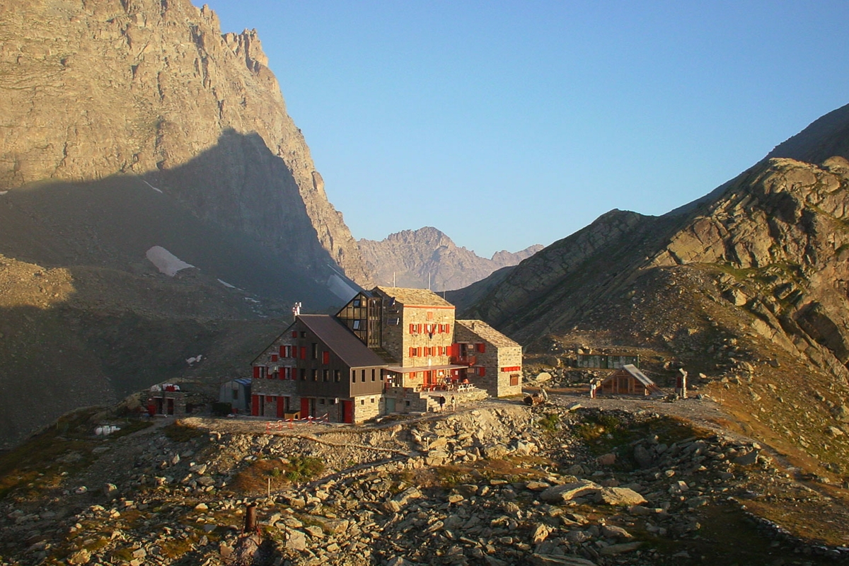 Foto del Rifugio Quintino Sella al Monviso: un edificio in tre corpi con imposte rosse, alle cui spalle si trova la mole del Monviso.