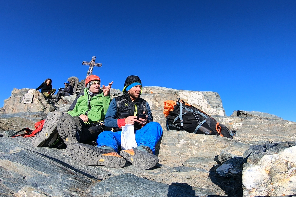 Paolo e Adriano, vestiti da montagna con giacca, caschetto e imbrago sono seduti sulle rocce poco al di sotto della Cima del Monviso, che si vede dietro di loro, dove sono seduti altri due scalatori