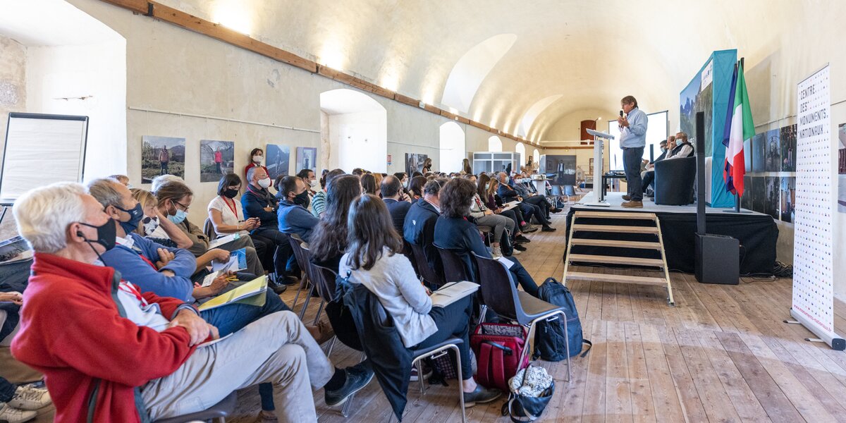In una lunga sala, all'interno di una cittadella fortificata, un folto pubblico seduto sta ascoltando un relatore. Il pubblico è a sinistra, il relatore su un podio a destra.