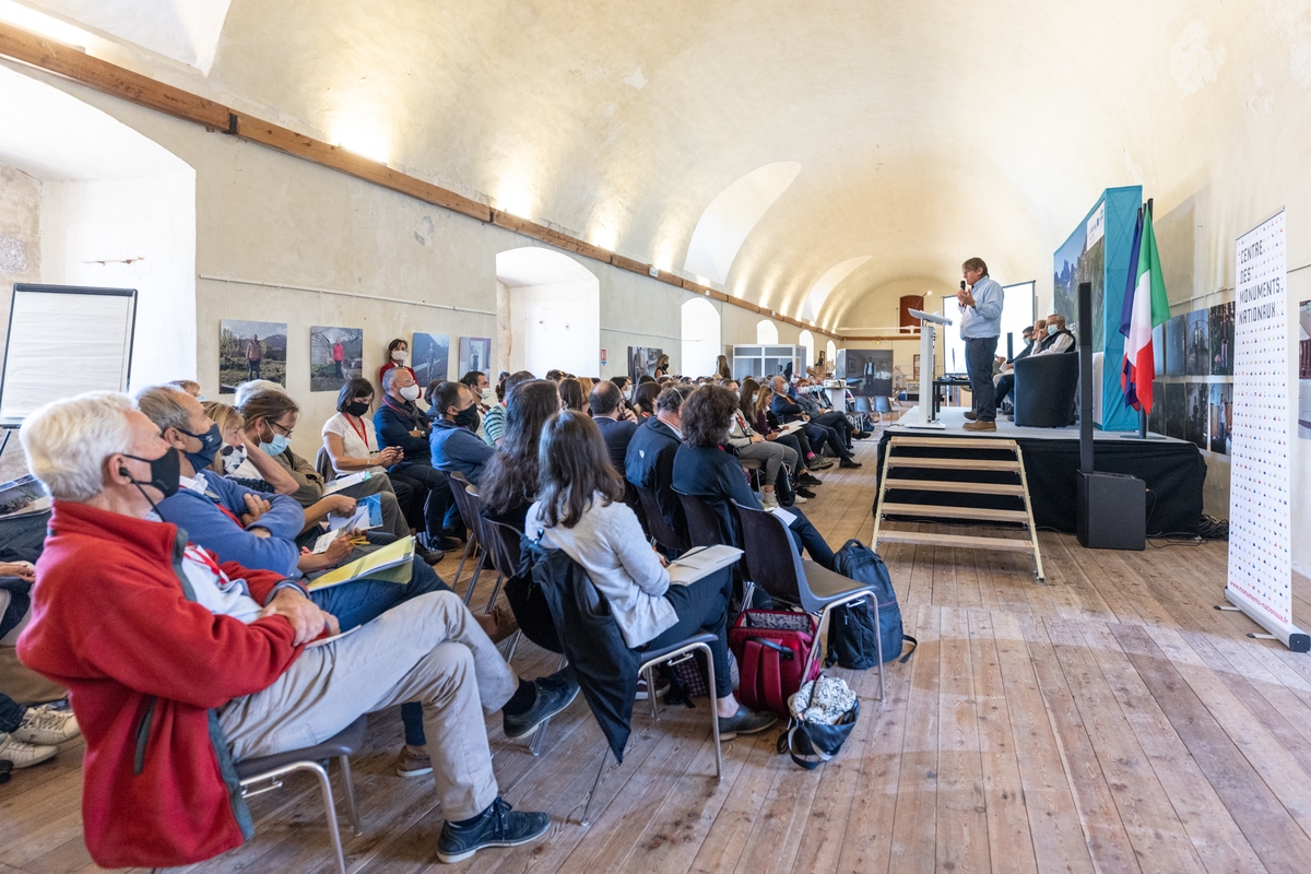 In una lunga sala, all'interno di una cittadella fortificata, un folto pubblico seduto sta ascoltando un relatore. Il pubblico è a sinistra, il relatore su un podio a destra.