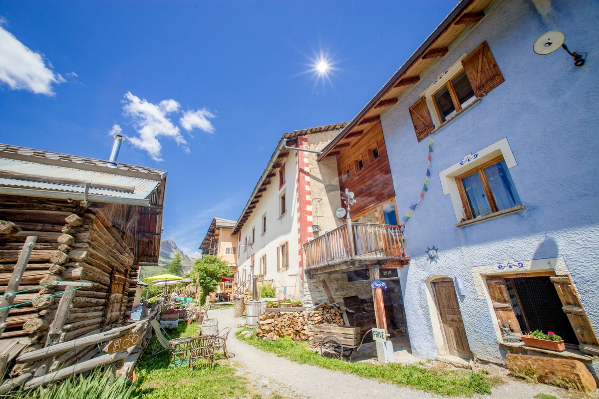 una serie di edifici con in mezzo una stradina sterrata. A sinistra una struttura in legno, a destra tre edifici, in primo piano uno azzurro, poi una struttura in legno con un terrazzino e quindi un edificio più grande. sulla strada un tavolini e sedie, una catasta di legna e un carretto. ambiente montano e cielo azzurro