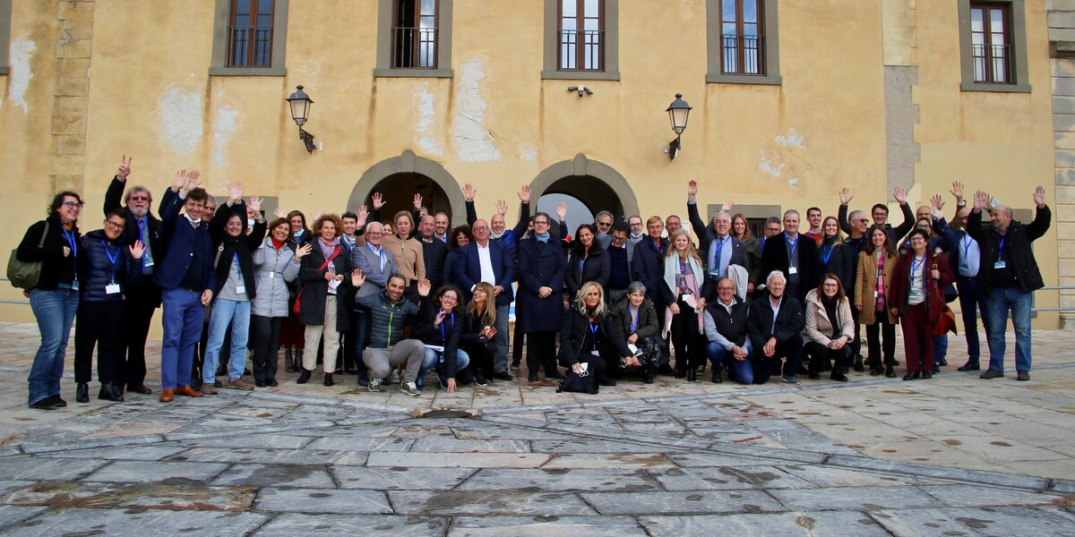 foto di gruppo: una cinquantina di persone tra donne e uomini riuniti su una piazza pavimentata in lastre di pietra; sullo sfondo un edificio giallo