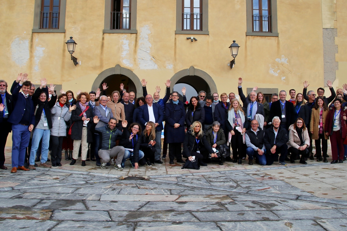 foto di gruppo: una cinquantina di persone tra donne e uomini riuniti su una piazza pavimentata in lastre di pietra; sullo sfondo un edificio giallo