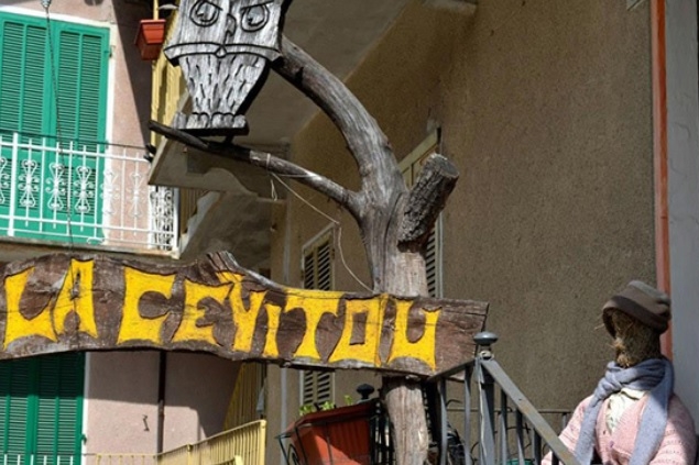 Scritta Cervitou intagliata nel legno e dipinta di giallo appesa a un tronco davanti a una casa. sul tronco anche un gufo intagliato nel legno. vicino alla casa uno spaventapasseri