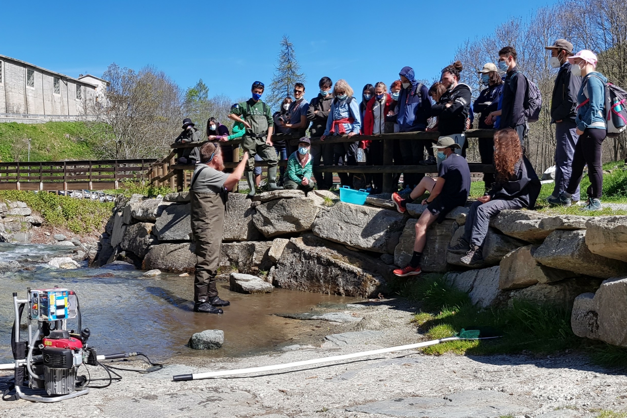 Il Prof. Fenoglio tiene una lezione a un gruppo di studenti durante una escursione didattica sul fiume Po, in località Crissolo. Si osserva il Prof. Fenoglio in acqua mentre mostra una trota appena catturata tramite elettronarcosi agli studenti, che sono disposti a semicerchio sulla riva.