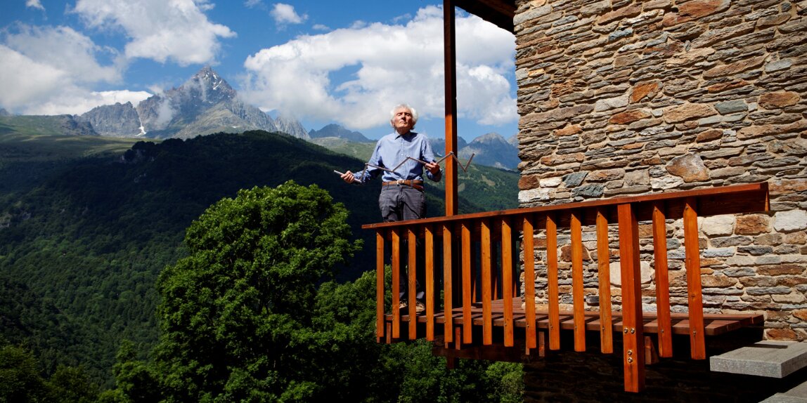 L'architetto Renato Maurino sul balcone di casa sua a Ostana: sullo sfondo c'è il Monviso, il balcone ha listelli di legno verticali come ringhiera.