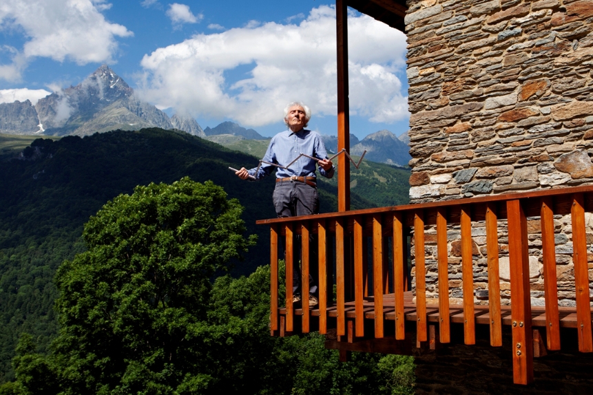L'architetto Renato Maurino sul balcone di casa sua a Ostana: sullo sfondo c'è il Monviso, il balcone ha listelli di legno verticali come ringhiera.