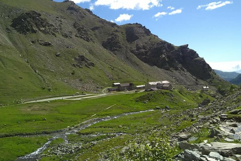 Il pianoro verde di Pian del Re a inizio giugno, in primo piano le rocce del sentiero, verso il centro i rigagnoli d'acqua che sgorgano dalla sorgente, al centro la strada e il rifugio albergo pian del re. intorno le montagne e cielo azzurro
