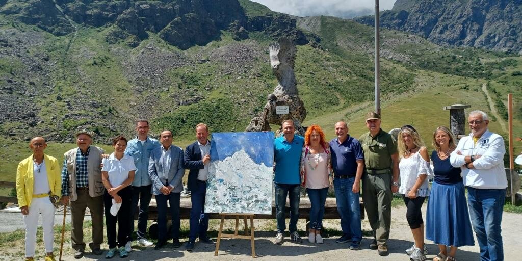 Un gruppo di persone ai due lati di un dipinto che raffigura il Monviso; sullo sfondo i rilievi dell'alta valle Po con un cielo coperto da nuvole