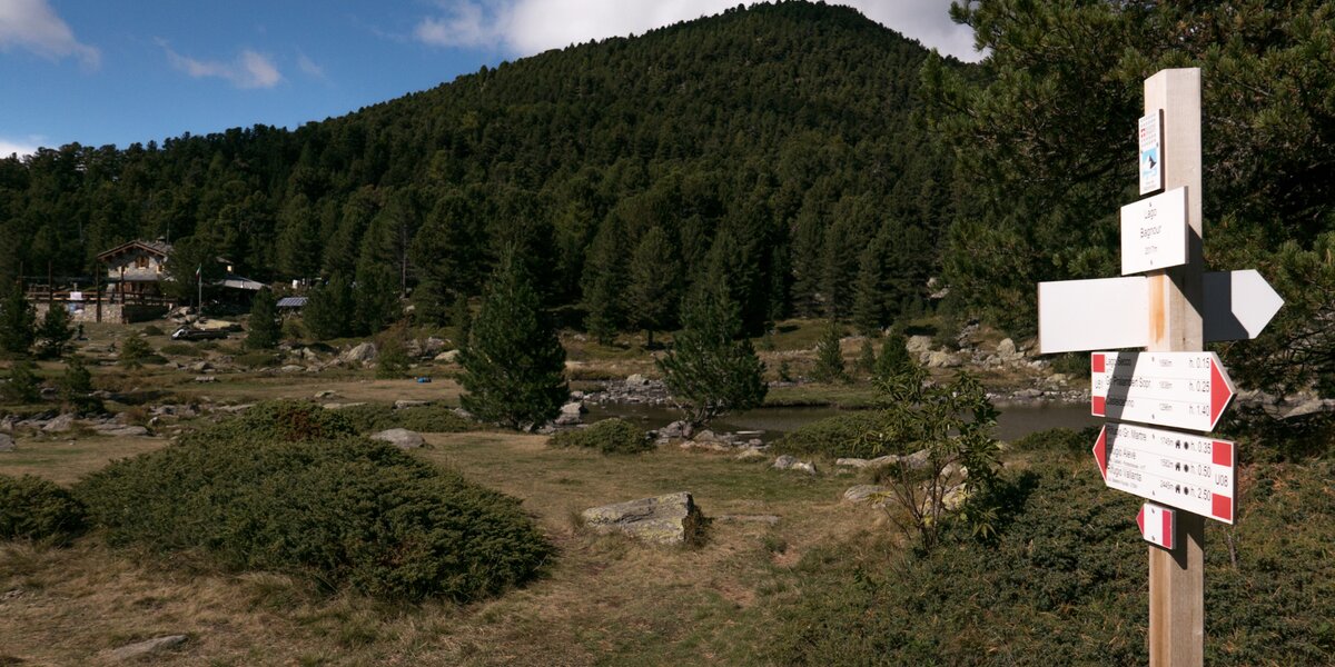 Una palina con cartelli indicatori e il logo del Parco del Monviso; sullo sfondo il lago Bagnour e il rifugio omonimo.