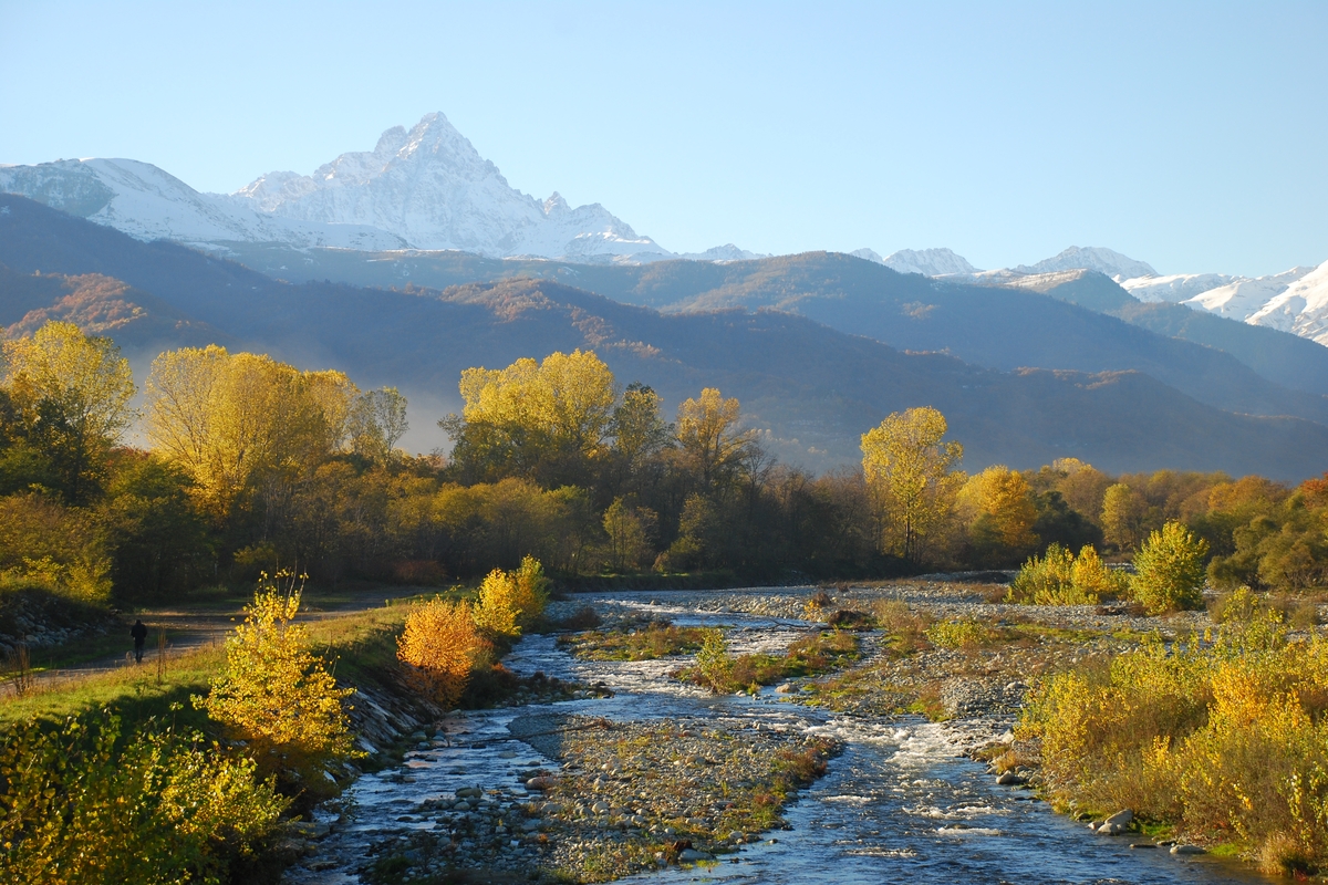 In primo piano il fiume Po su un greto di ciottoli e pietre, sullo sfondo il Monviso, intorno alberi in veste autunnale con colori gialli e marroni
