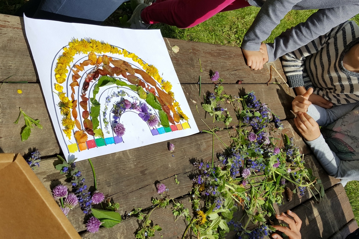 un arcobaleno realizzato su un foglio con dei fiori. il foglio è appoggiato su un tavolo dove sono presenti altri petali. si vedono le mani di bambini appoggiati sul tavolo