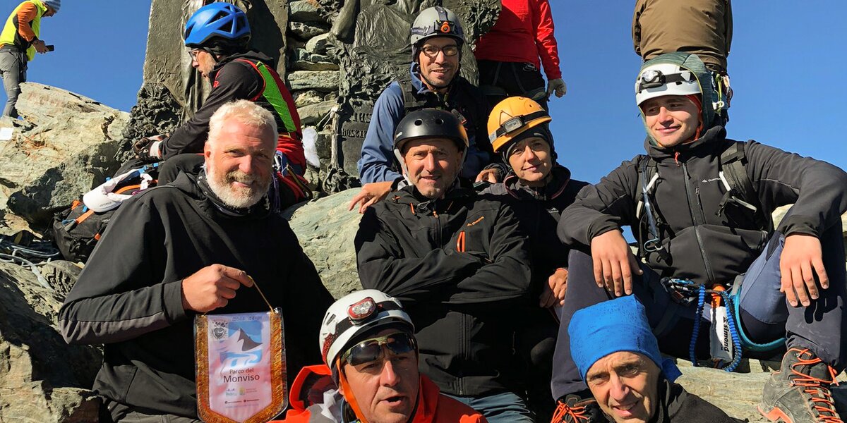 un gruppo di persone in abbigliamento da alta montagna sulla vetta del Monviso. Una persona, con barba e capelli bianchi posta sulla sinistra dell'immagine, regge e mostra un gagliardetto con lo stemma del Parco del Monviso.