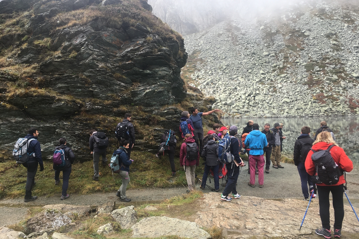 un gruppo di persone guarda le rocce adiacenti al lago Fiorenza, visibile sulla parte destra dell'immagine. La giornata è nebbiosa e tutti indossano giacche a vento.