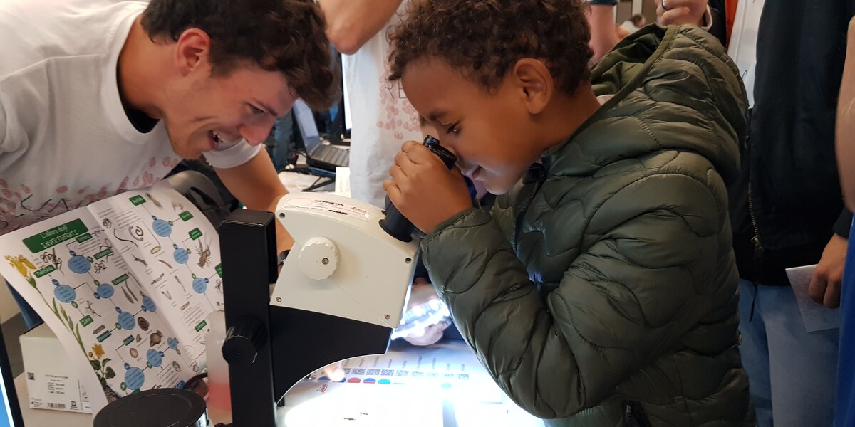 un bambino osserva in uno stereoscopio alcuni macroinvertebrati, mentre un tesiste del gruppo Alpstream lo guida nell'osservazione dei particolori morfologici
