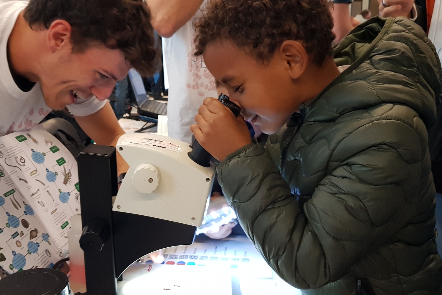 un bambino osserva in uno stereoscopio alcuni macroinvertebrati, mentre un tesiste del gruppo Alpstream lo guida nell'osservazione dei particolori morfologici