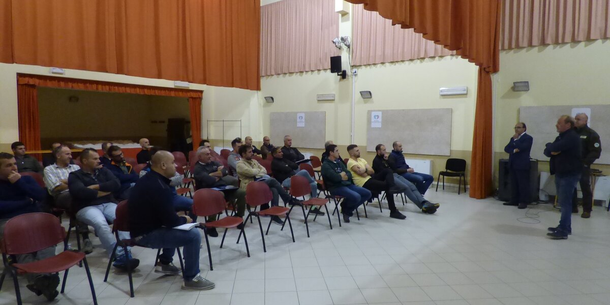 a destra una persona sta parlando davanti ad una platea di circa trenta persone, sulla sinistra dell'immagine.