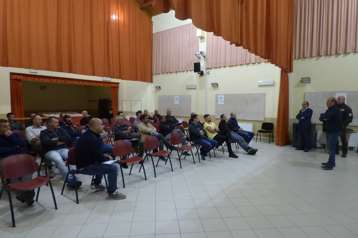 a destra una persona sta parlando davanti ad una platea di circa trenta persone, sulla sinistra dell'immagine.