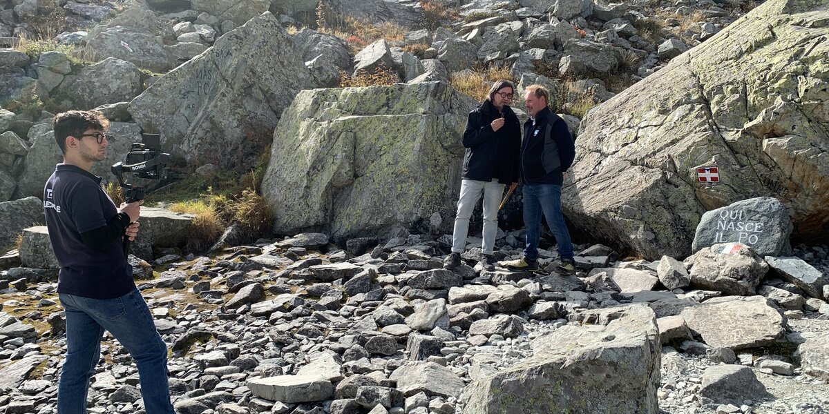 Alle sorgenti del Po, visibili sulla destra dell'immagine, un cameraman sta inquadrando due persone che parlano: il conduttore Claudio Calorio e il presidente del Parco del Monviso Dario Miretti.