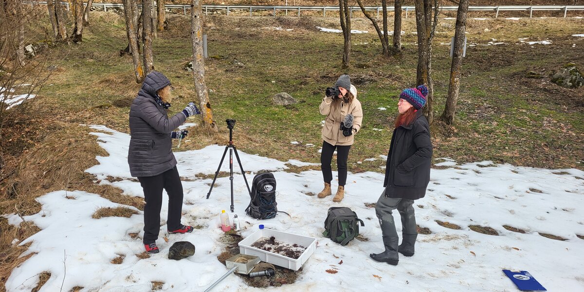 Tre ricercatrici stanno lavorando all'aperto, in un prato parzialmente ricoperto di neve: stanno effettuando alcune riprese video del progetto in corso di svolgimento.