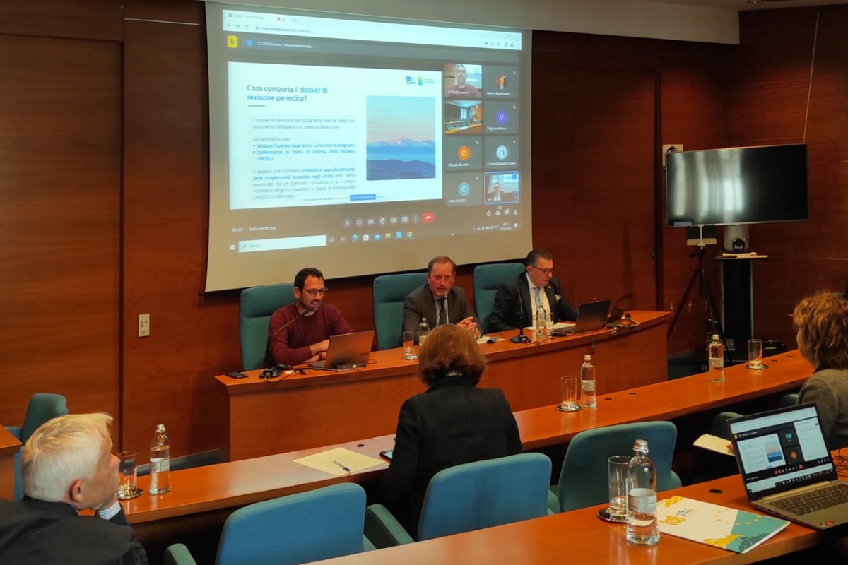 In una sala rivestita da pannellature in legno si vede al centro della foto un tavolo dei relatori con tre persone al di sopra delle quali uno schermo sta proiettando le immagini di una presentazione