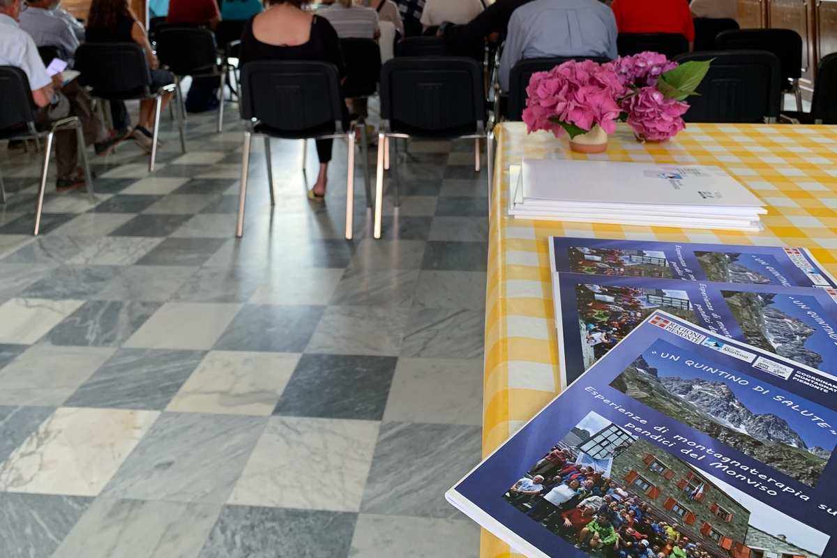 in primo piano, sulla destra, opuscoli informativi su Officina Monviso: sullo sfondo si vedono le schiene dei partecipanti alla conferenza di presentazione dell'edizione 2023 di Officina Monviso.
