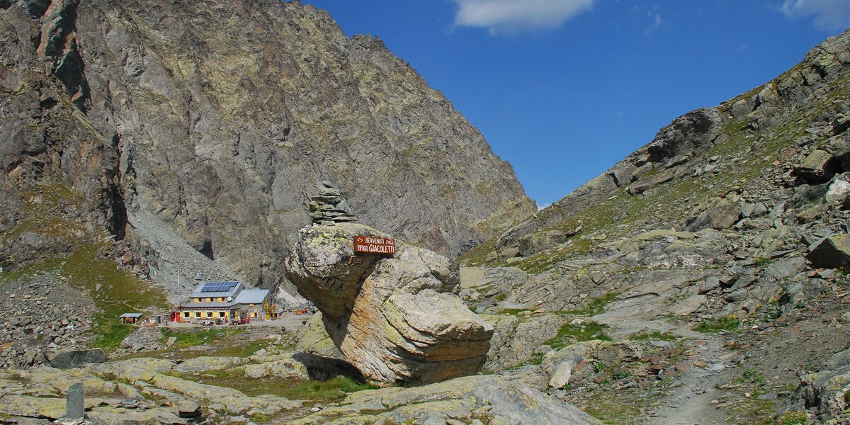 in primo piano una pietra con un cartello di legno di benvenuto al rifugio, dietro il rifugio con le persiane gialle, sullo sfondo montagne e cielo azzurro
