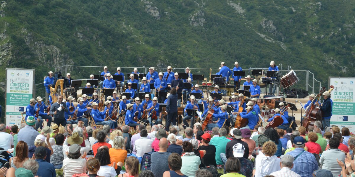 foto di repertorio della scorsa edizione del Concerto di Ferragosto a Paesana, del 2018: nella parte bassa dell'immmagine il pubblico di schiena, seduto per terra, in alto l'orchestra intenta a suonare.