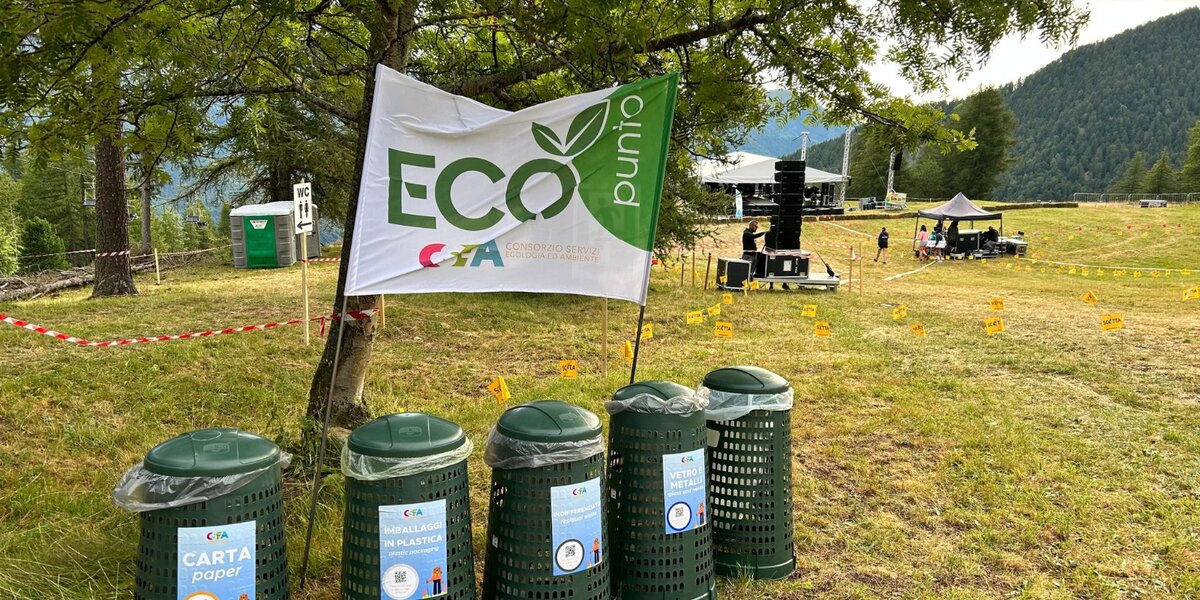 Cinque contenitori per rifiuti differenziati in plastica verde in un prato; sullo sfondo si vede il palco di un evento musicale in corso di organizzazione (foto di repertorio)