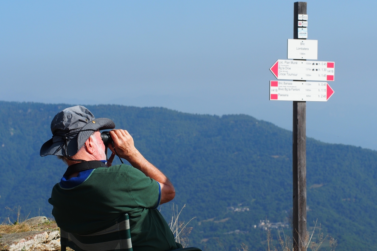 Un uomo di spalle, seduto e con un binocolo, è rivolto verso il fondovalle dal Bric Lombatera di Paesana: si vede il cartello indicatore della località, posta a quota 1.390 metri in valle Po
