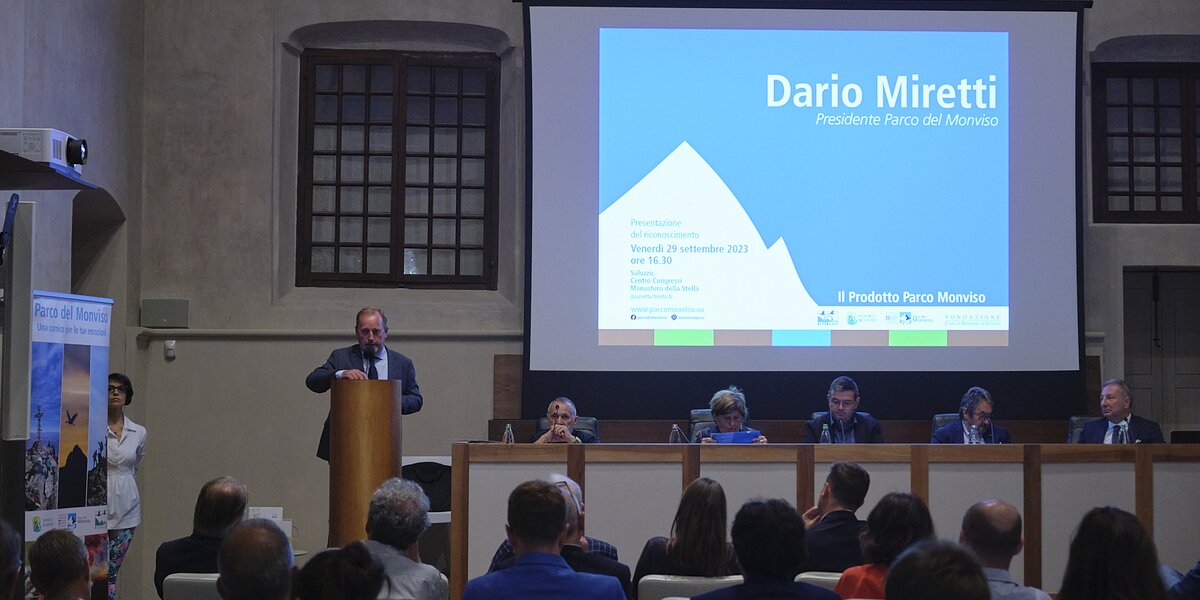 Un momento della presentazione del riconoscimento Prodotto Parco Monviso: sta parlando il presidente Dario Miretti che si trova al un leggio, alla sinistra dell'immagine. Sulla destra uno schermo proietta il suo nome e al di sotto c'è un tavolo relatori con cinque persone. Nella parte bassa della fotografia si vede il pubblico di spalle