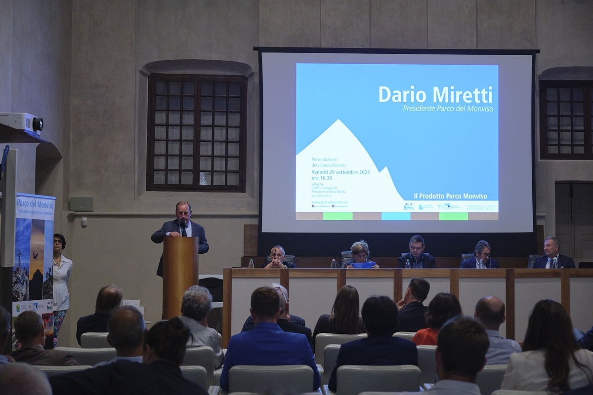 Un momento della presentazione del riconoscimento Prodotto Parco Monviso: sta parlando il presidente Dario Miretti che si trova al un leggio, alla sinistra dell'immagine. Sulla destra uno schermo proietta il suo nome e al di sotto c'è un tavolo relatori con cinque persone. Nella parte bassa della fotografia si vede il pubblico di spalle