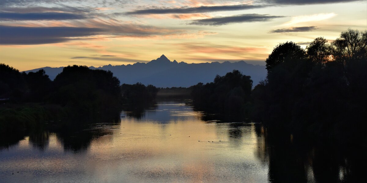 Il Monviso al centro della foto, in alto, si specchia sul fiume Po al tramonto. L'immagine ha una dominante di colore blu.
