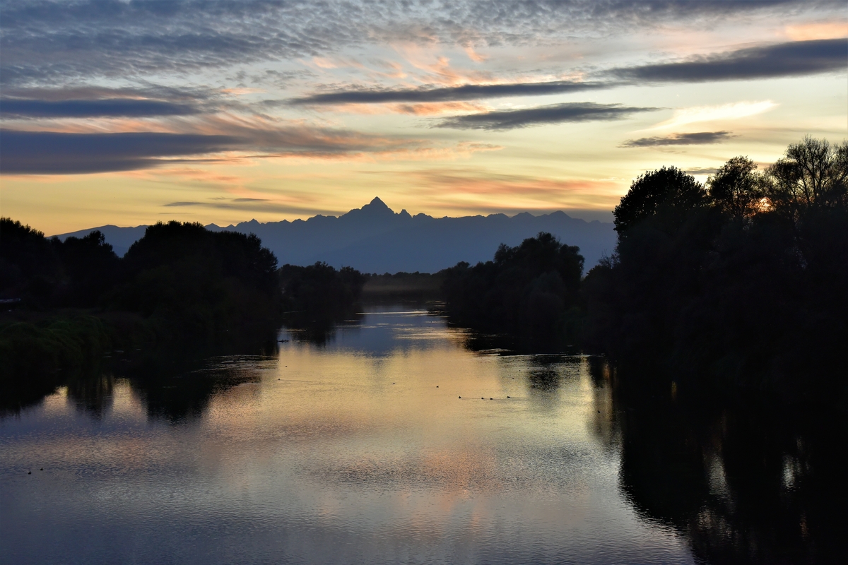 Il Monviso al centro della foto, in alto, si specchia sul fiume Po al tramonto. L'immagine ha una dominante di colore blu.