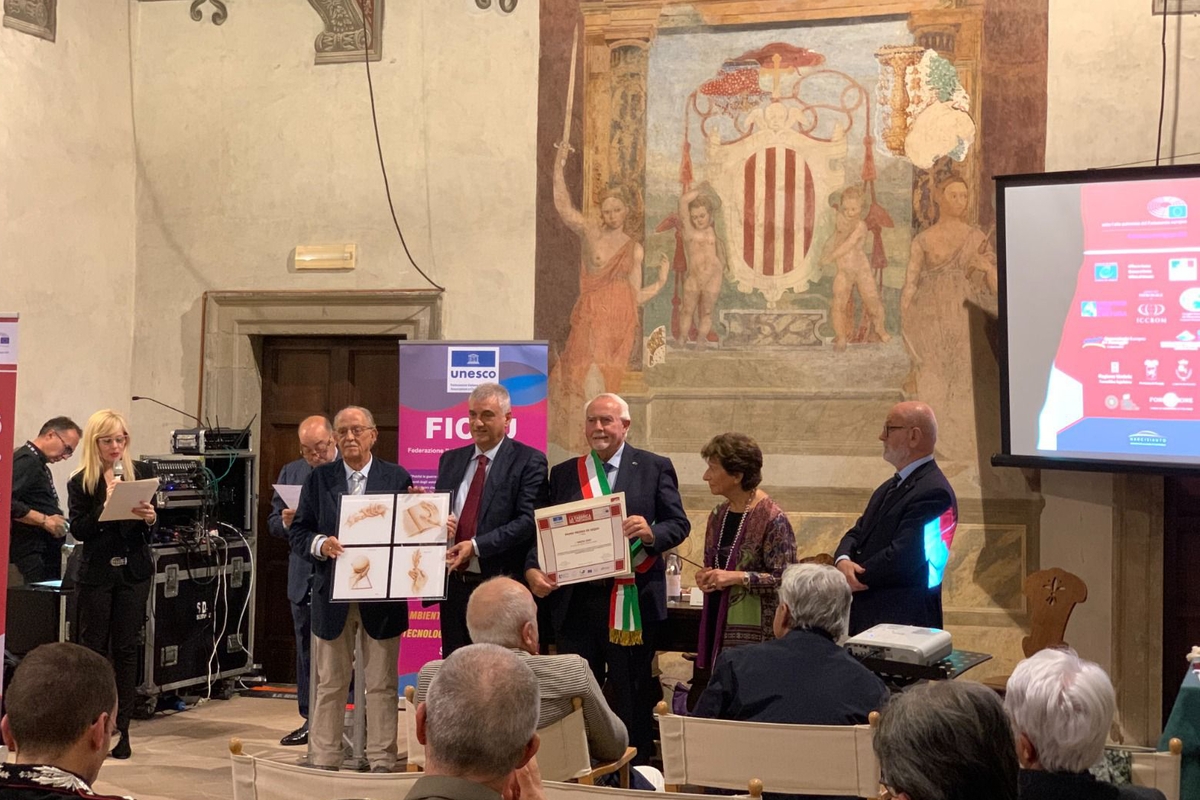 in una sala riccamente affrescata, alcune persone di schiena sono il pubblico della premiazione del Sindaco di Macra, che è posto al centro della foto e sta ricevendo una pergamena dalle mani di due persone. Intorno ad essi, altre figure.