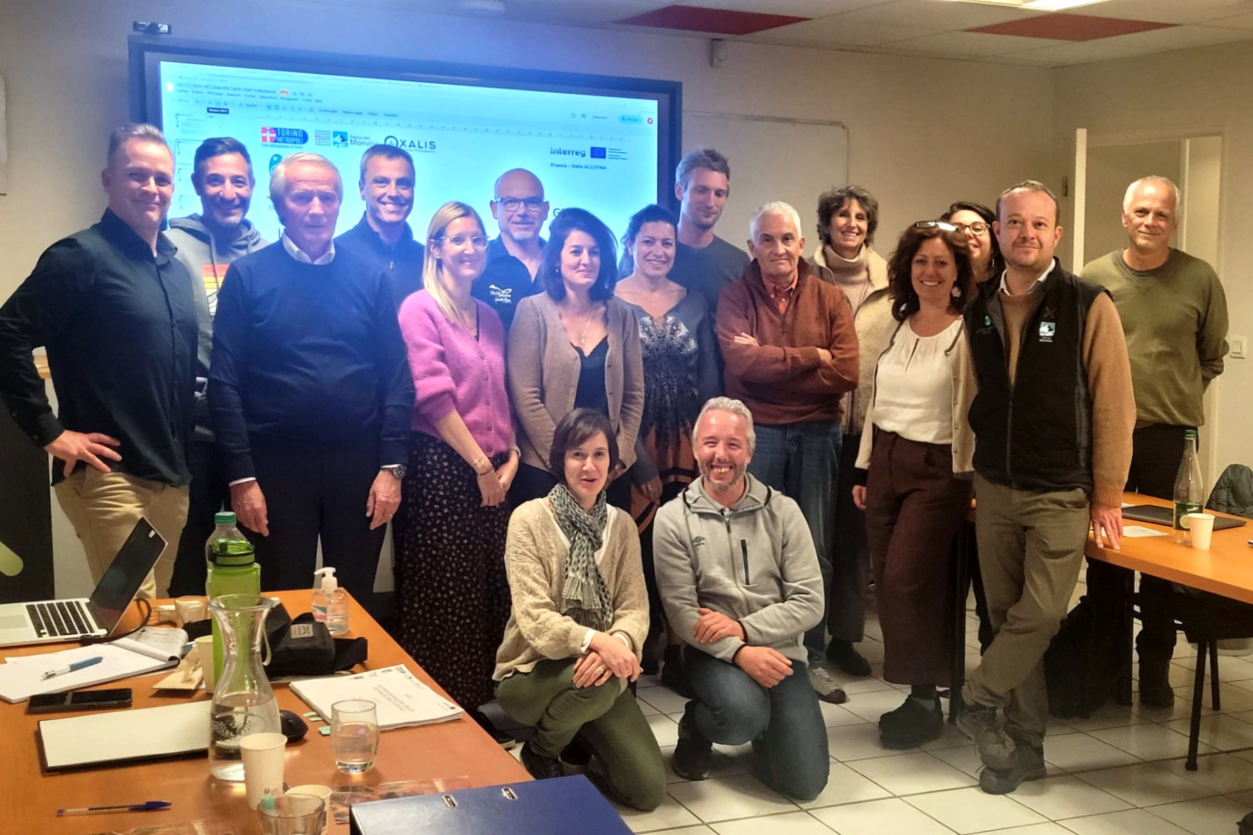 foto di gruppo dei partecipanti alla riunione: 15 persone in piedi e 2 accucciate; dietro di loro una lavagna luminosa. Davanti a sinistra, una scrivania con computer e altri oggetti sopra.