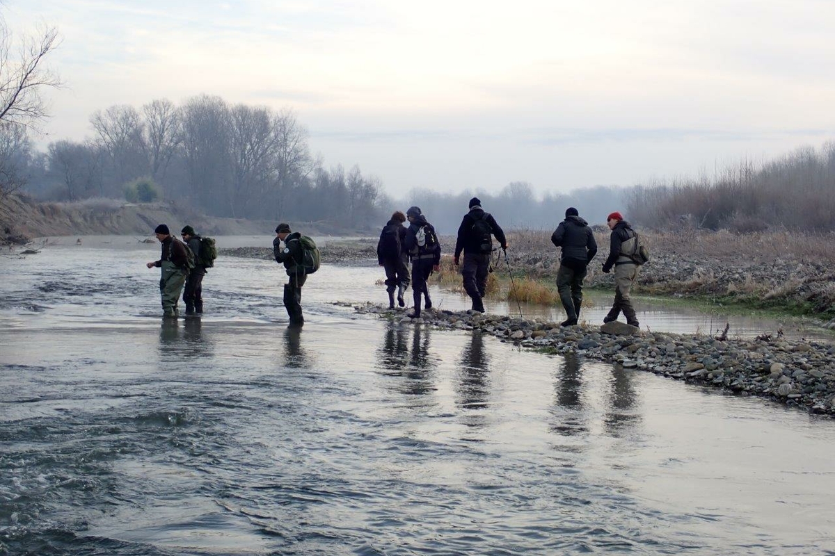 Al centro della foto si vedono otto persone di schiena che si trovano nell'alveo di un corso d'acqua: si tratta dei tecnici impegnati nel monitoraggio. Intorno a loro si vedono le rive del fiume e una scarna vegetazione invernale, sotto ad un cielo grigio
