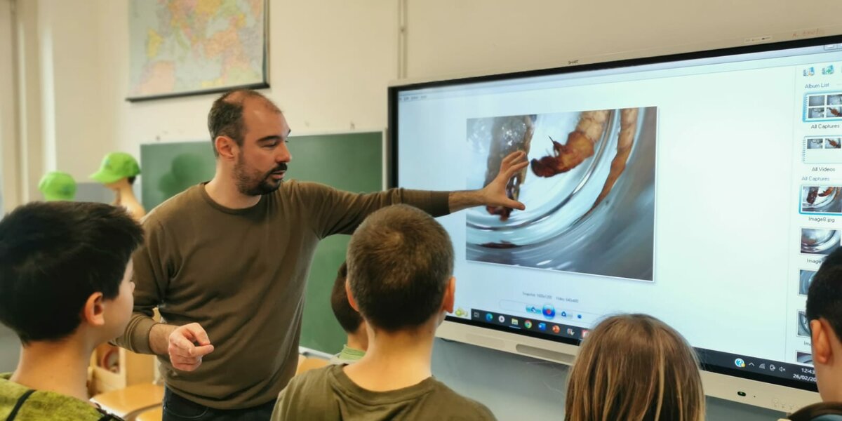 Il ricercatore mostra con la mano un'immagine sulla lavagna luminosa della classe, con quattro studenti di spalle che guardano da vicino
