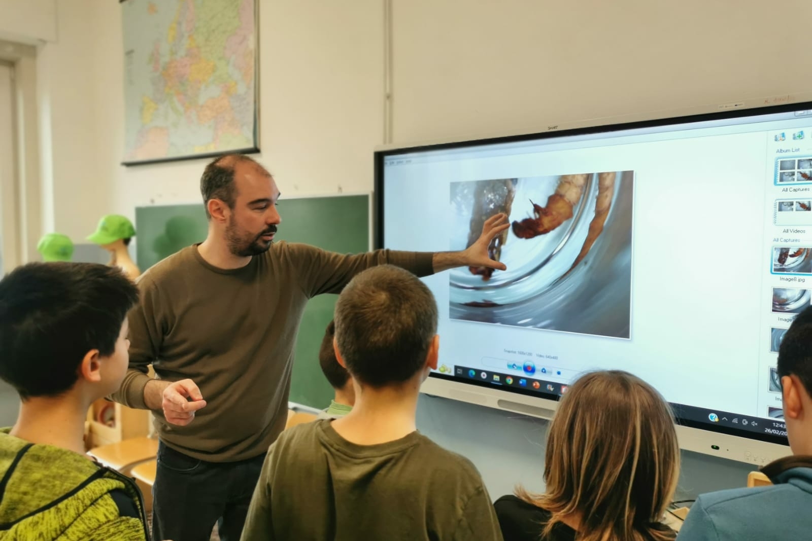 Il ricercatore mostra con la mano un'immagine sulla lavagna luminosa della classe, con quattro studenti di spalle che guardano da vicino
