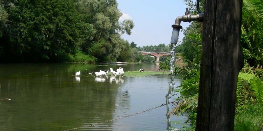 Una fontana in primo piano con sullo sfondo il fiume Po nel quale si trovano alcune oche; ancora più in dietro un ponte.