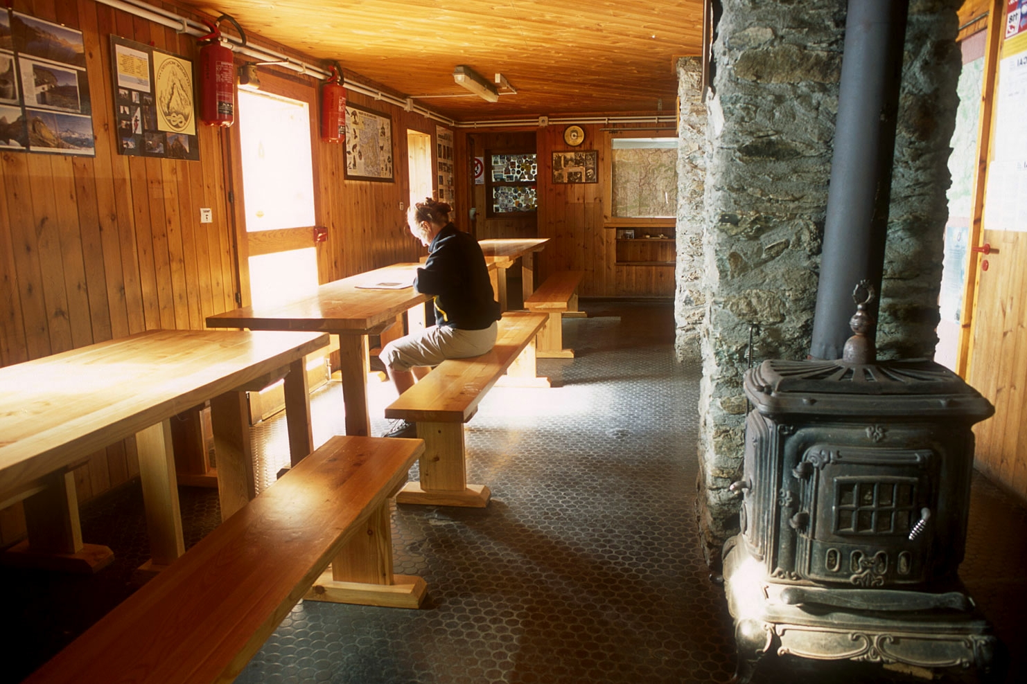 Una persona seduta ad una tavolata all'interno del rifugio: a destra in primo piano una grande stufa