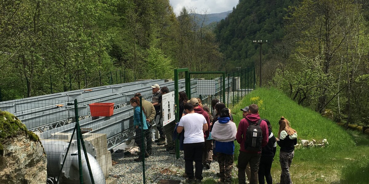 un gruppo di persone di spalle sta entrando in un'area recintata dove si trovano tre canalette in metallo, usate a scopo di studio sugli ecosistemi fluviali.