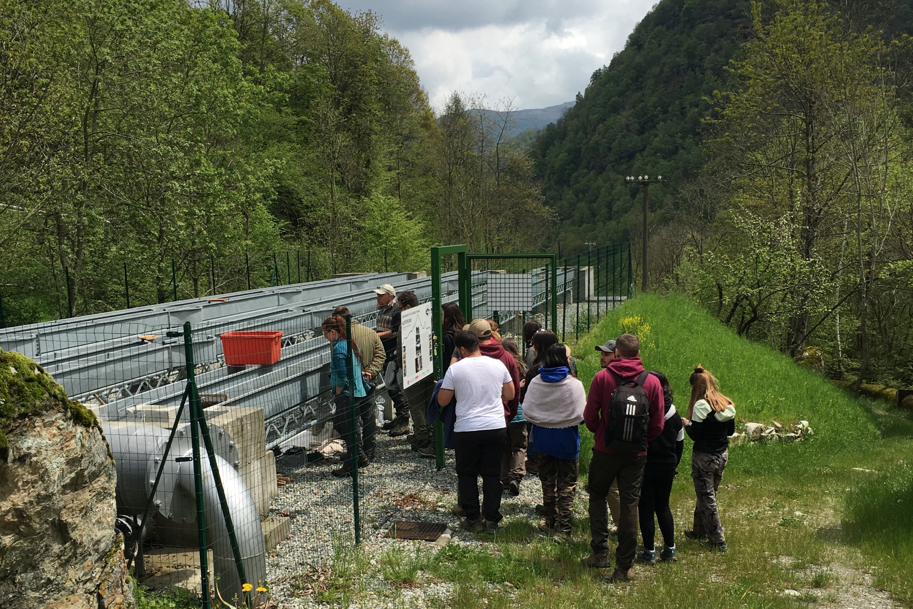 un gruppo di persone di spalle sta entrando in un'area recintata dove si trovano tre canalette in metallo, usate a scopo di studio sugli ecosistemi fluviali.