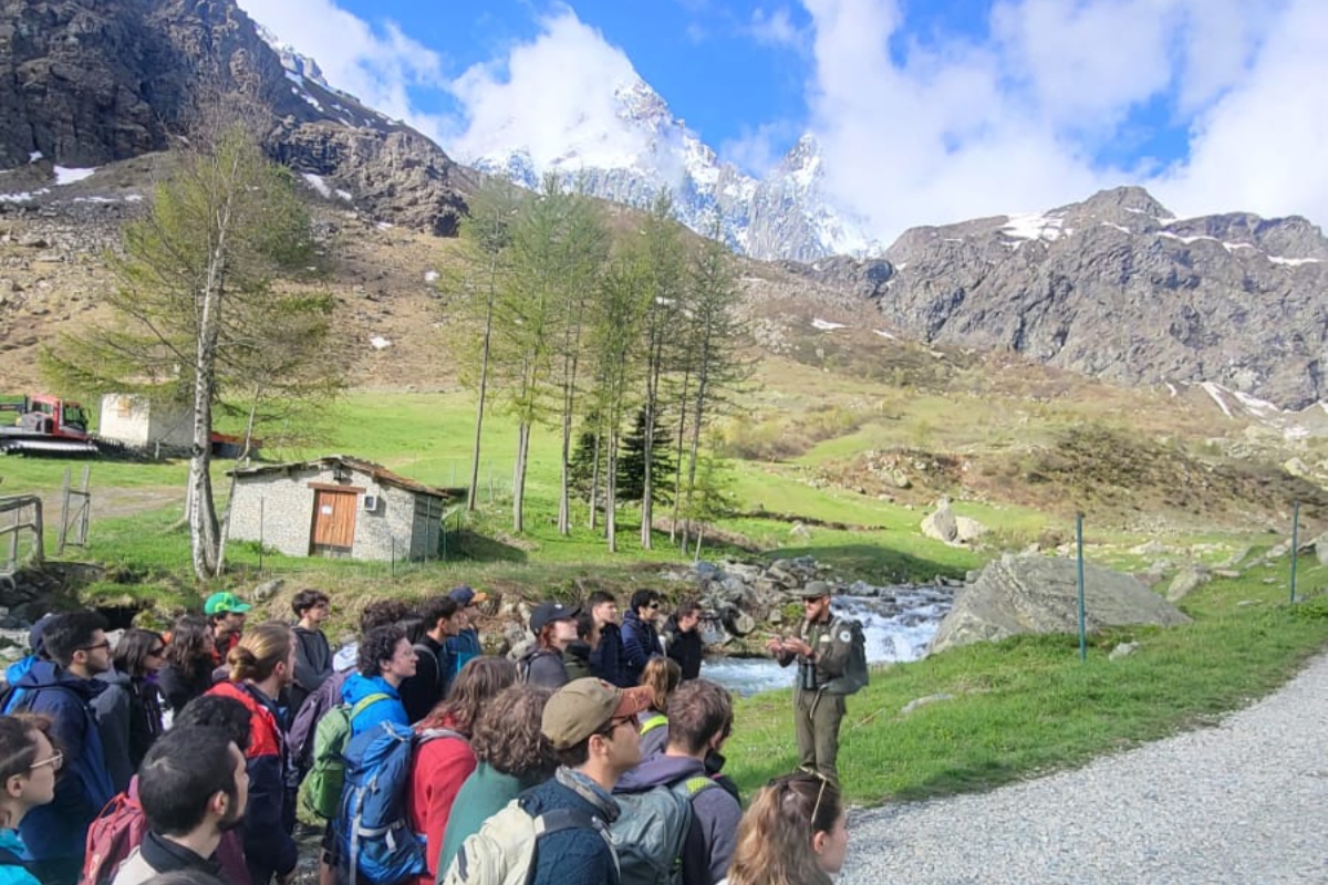in basso a sinistra un gruppo di studenti che sta ascoltando un guardiaparco all'aperto, nei pressi del fiume Po e al cospetto del Monviso, che si intravede al centro della fotografia in alto, coperto parzialmente da qualche nuvola.