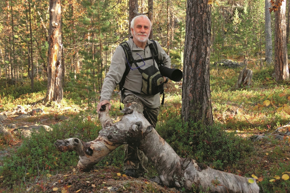 un uomo in un bosco, vestito con abiti da escursione e con materiale fotografico, si appoggia ad un tronco curvo caduto a terra.