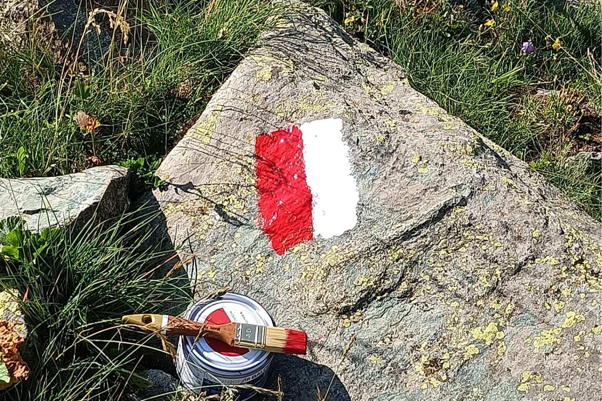 una tacca segnavia in vernice bianca e rossa su una roccia, a lato il pennello e la vernice usata per ridipingere la traccia