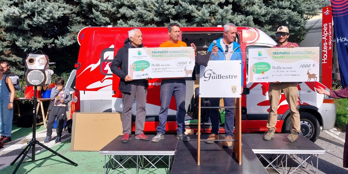 la consegna del premio: su un piccolo palco in strada una persona consegna due assegni di grandi dimensioni ai referenti dei progetti premiati