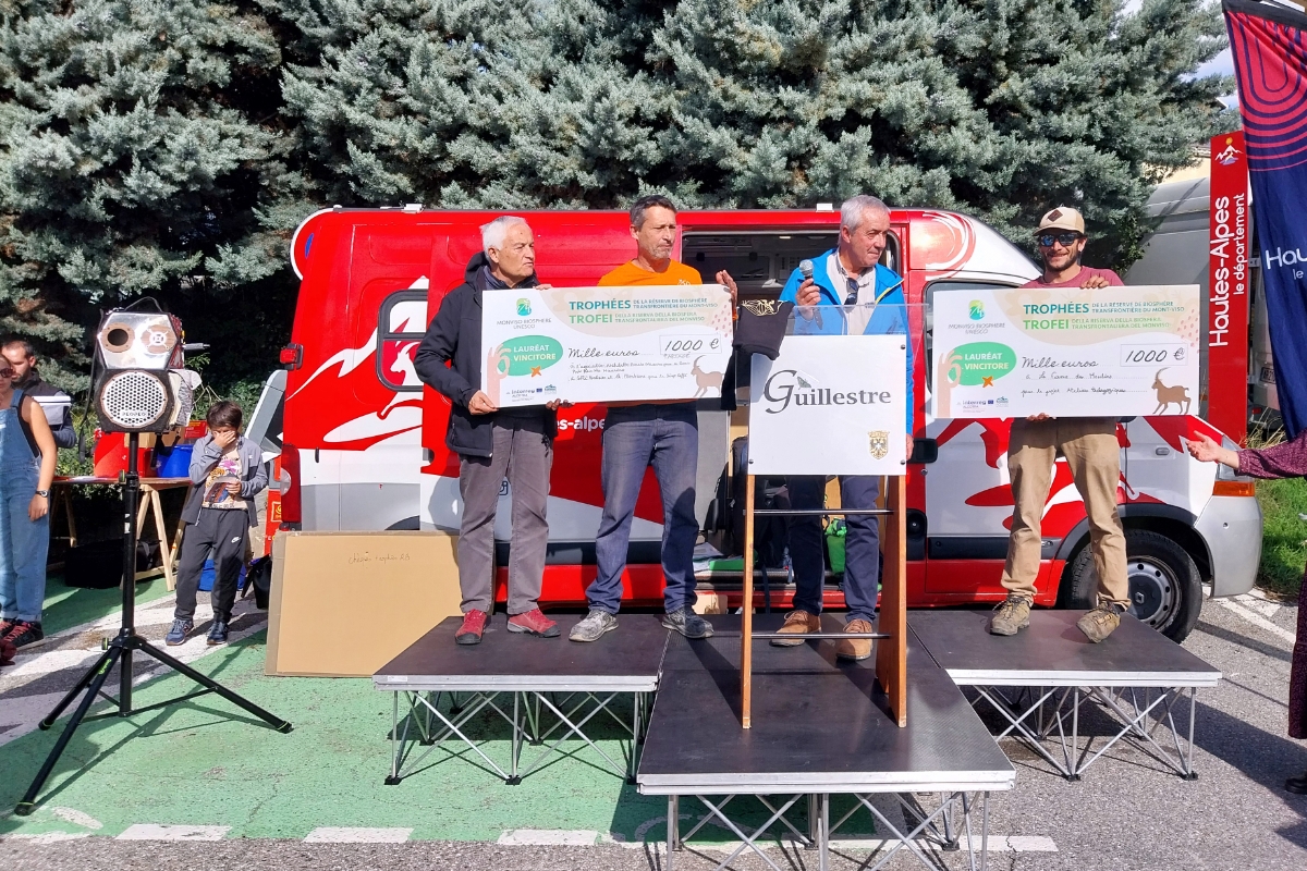 la consegna del premio: su un piccolo palco in strada una persona consegna due assegni di grandi dimensioni ai referenti dei progetti premiati